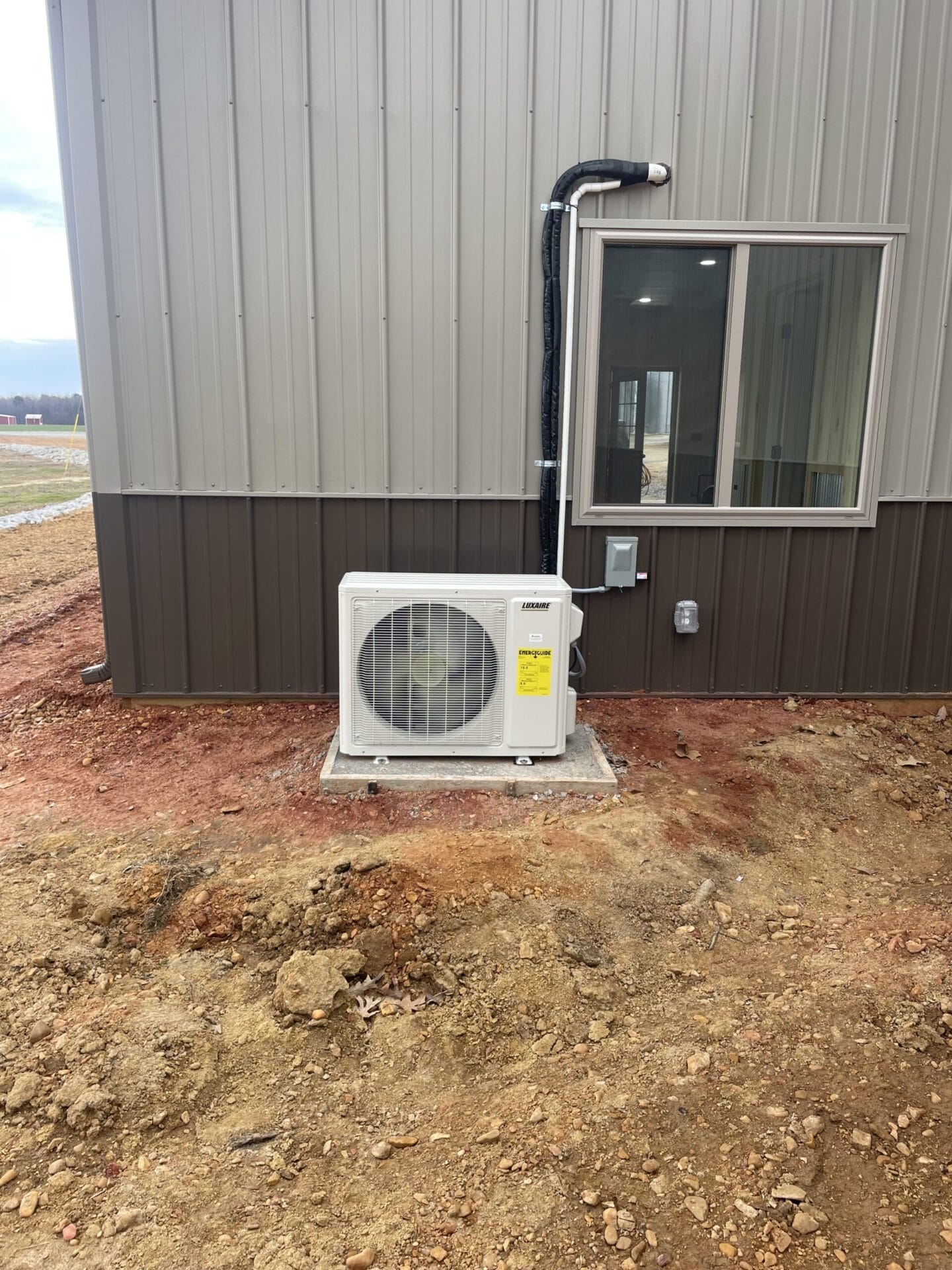 Air conditioning unit installed on a concrete pad outside a brown and gray building with a window.