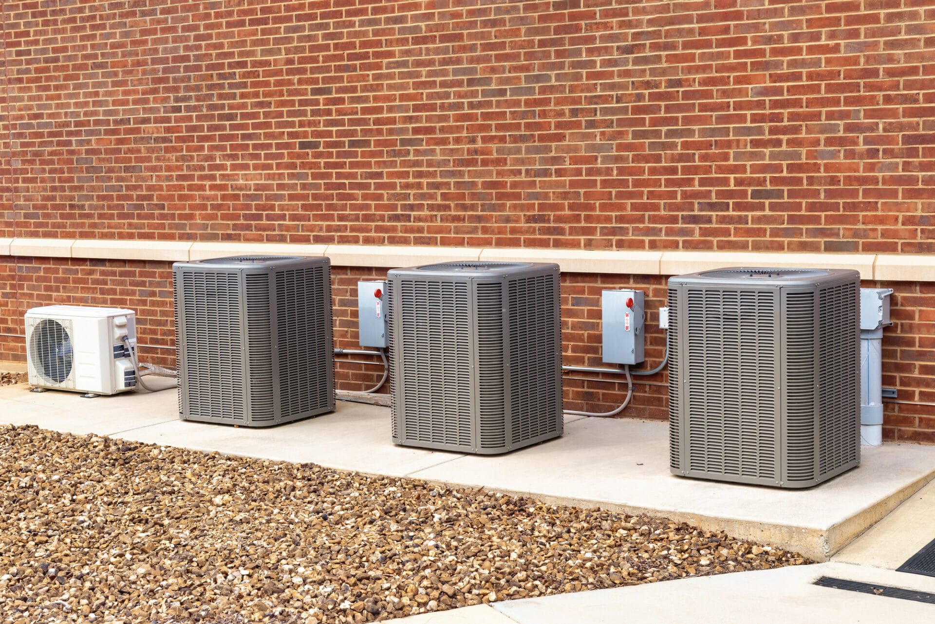 Three large air conditioning units and one smaller unit are mounted on a concrete pad against a brick wall.