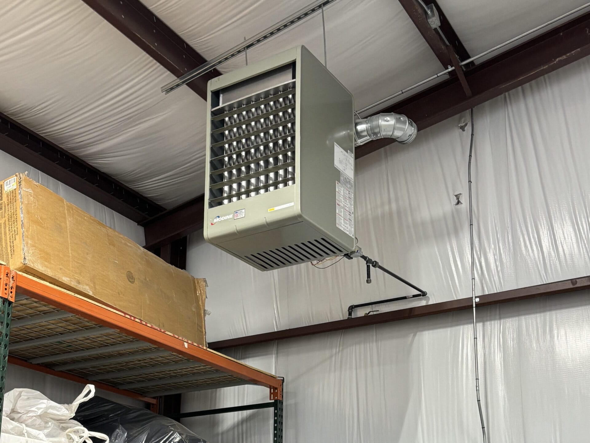 A ceiling-mounted industrial heater in a warehouse with insulation on the walls. A cardboard box is partially visible on the shelf below.