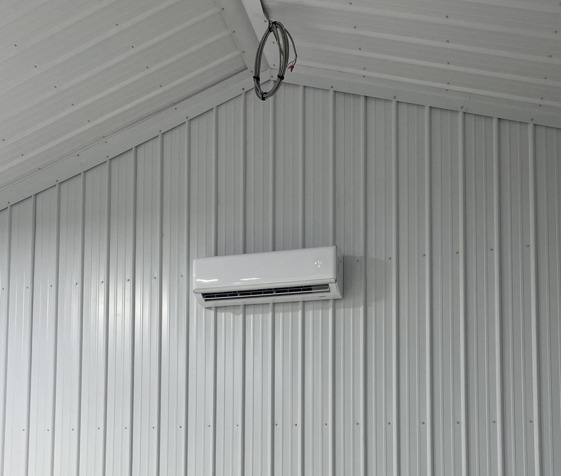 Wall-mounted air conditioning unit on a white corrugated metal wall inside a building with a corrugated metal ceiling.