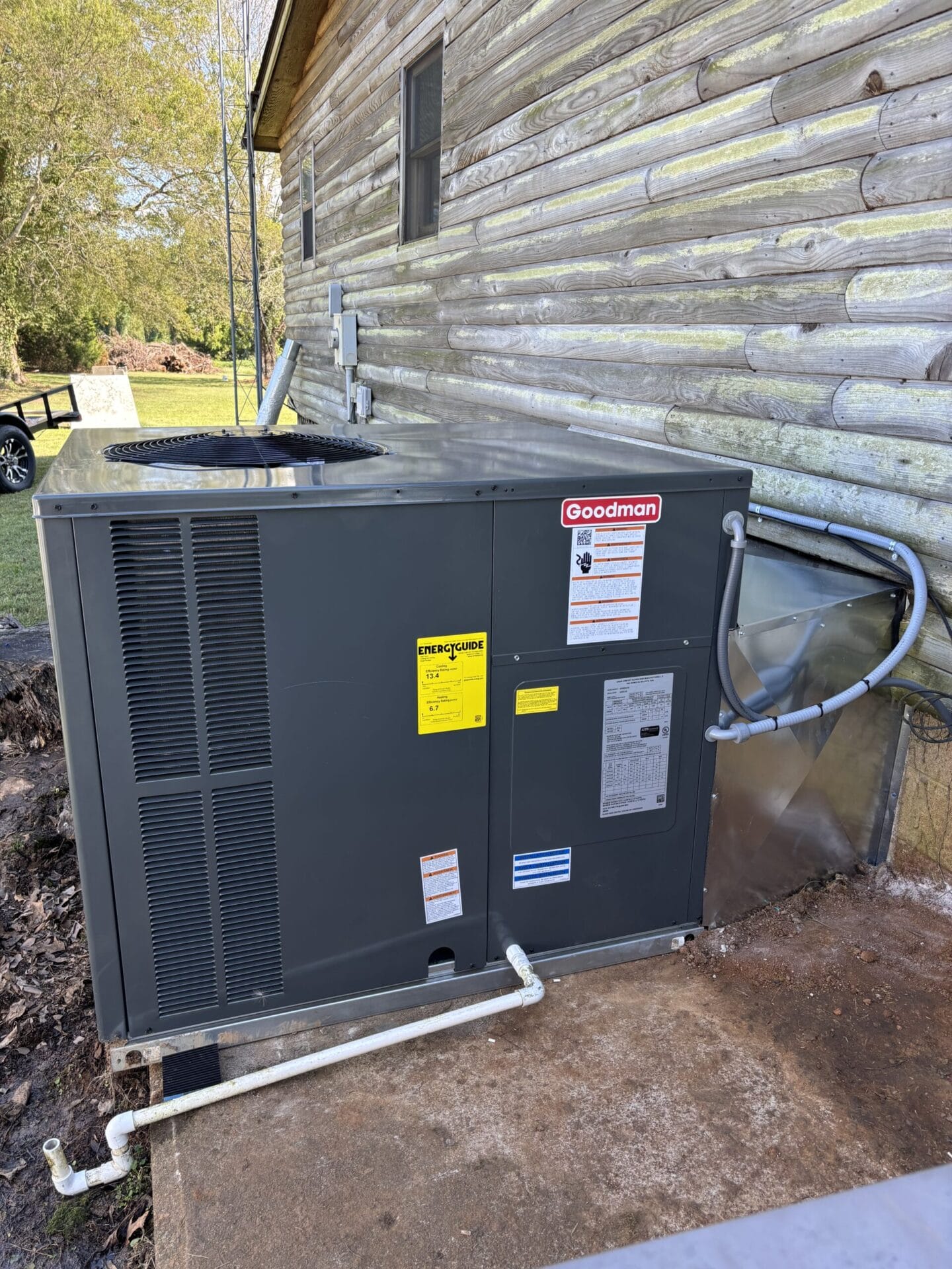 Outdoor HVAC unit installed next to a wooden house, with visible piping and electrical connections.