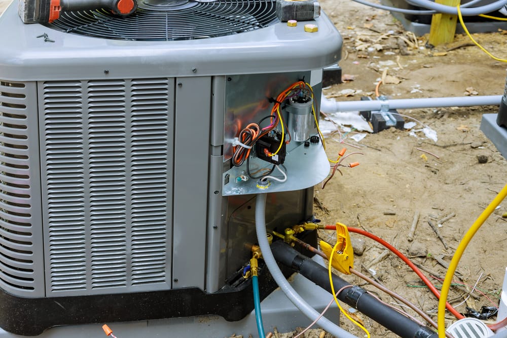 An outdoor heating unit with its side panel open, revealing internal wiring and components. Various tools and cables are visible around it.